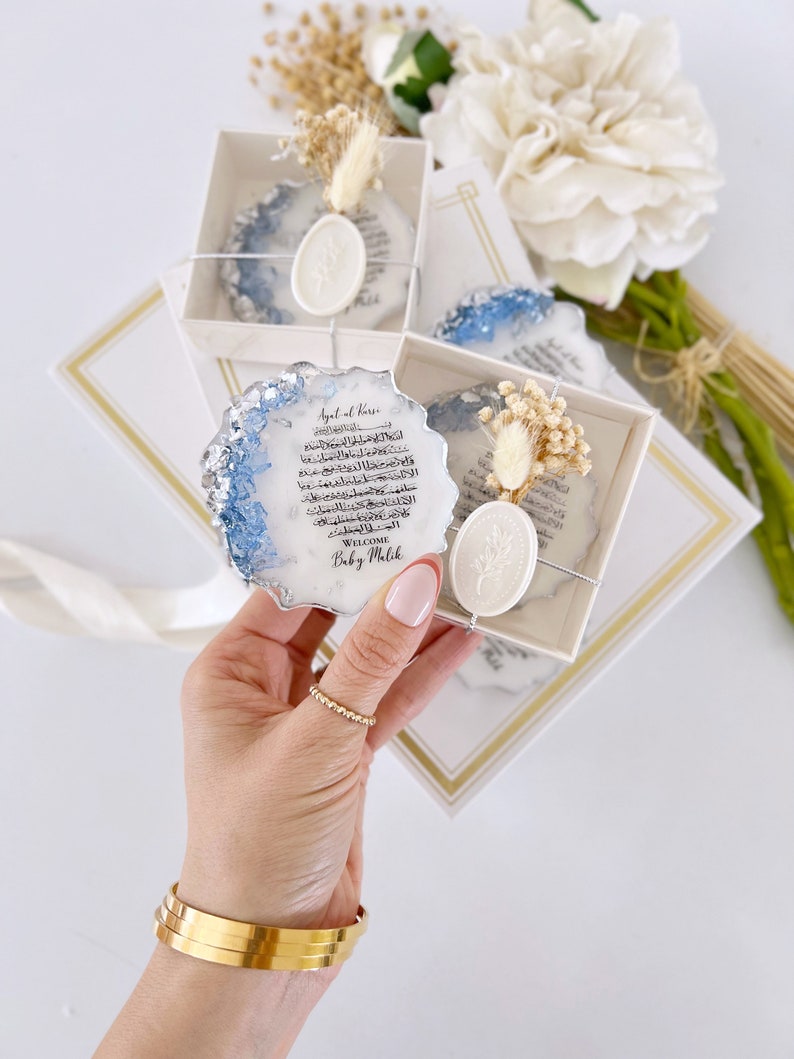 May include: A white box with a blue and silver resin coaster featuring Arabic calligraphy and a wax seal. The coaster is decorated with dried flowers and the text "Welcome Baby Malik".