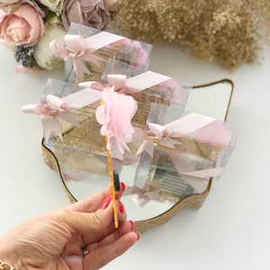 May include: A set of small, clear gift boxes with pink ribbon and a pink flower on top. The boxes are arranged on a gold-colored tray with a mirror surface. The boxes are filled with small, decorative items.