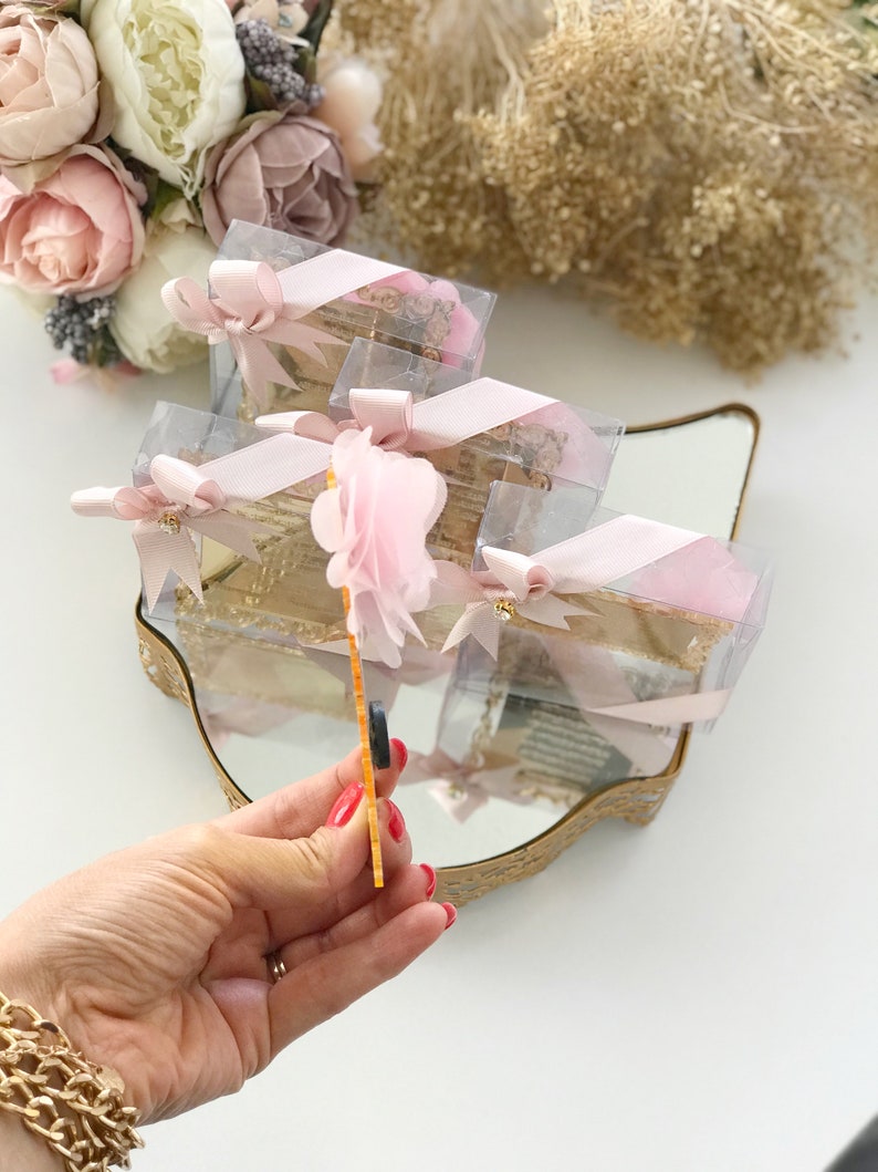 May include: A set of small, clear gift boxes with pink ribbon and a pink flower on top. The boxes are arranged on a gold-colored tray with a mirror surface. The boxes are filled with small, decorative items.
