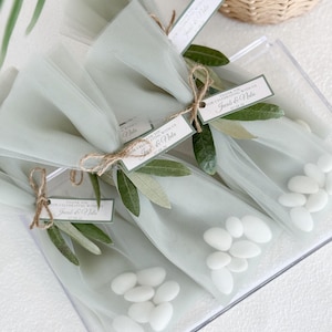May include: Clear plastic box containing several small, tulle-wrapped packages of white candies. Each package is tied with twine and adorned with olive branches and a tag that reads "Thank you for celebrating with us, Jacob & Nola."