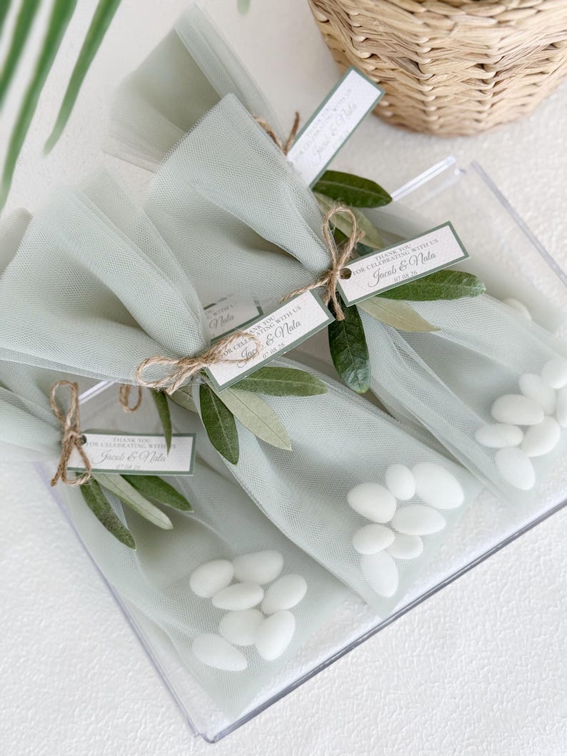 May include: Clear plastic box containing several small, tulle-wrapped packages of white candies. Each package is tied with twine and adorned with olive branches and a tag that reads "Thank you for celebrating with us, Jacob & Nola."