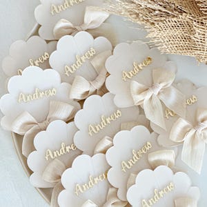 Puede incluir: Tarjetas de lugar con bordes festoneados y el nombre "Andrew" en escritura dorada. Cada tarjeta está adornada con un lazo de cinta color crema. Las tarjetas están dispuestas sobre un plato blanco, ideal para bodas o eventos.