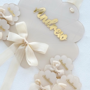 Op de afbeelding: Crèmekleurige naamkaartjes met geschulpte randen met de naam "Andrew" in gouden letters. Elk kaartje is versierd met een crèmekleurige strik. Een grotere, waaiervormige kaart met de naam "Andrew" is ook aanwezig, samen met crèmekleurige linten.