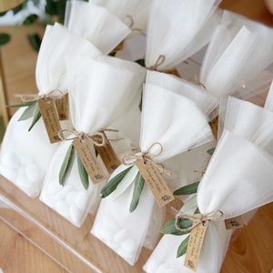 May include: Several small white tulle bags filled with white candies, tied with twine and adorned with olive branches and tags. The tags read "The Wedding of [Names]" and a date. The bags are arranged on a wooden surface.