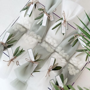 May include: Clear cellophane bags filled with white candies, tied with twine and adorned with olive branches and tags. The bags are arranged in a clear container, creating a visually appealing presentation. A perfect gift for a wedding or special event.