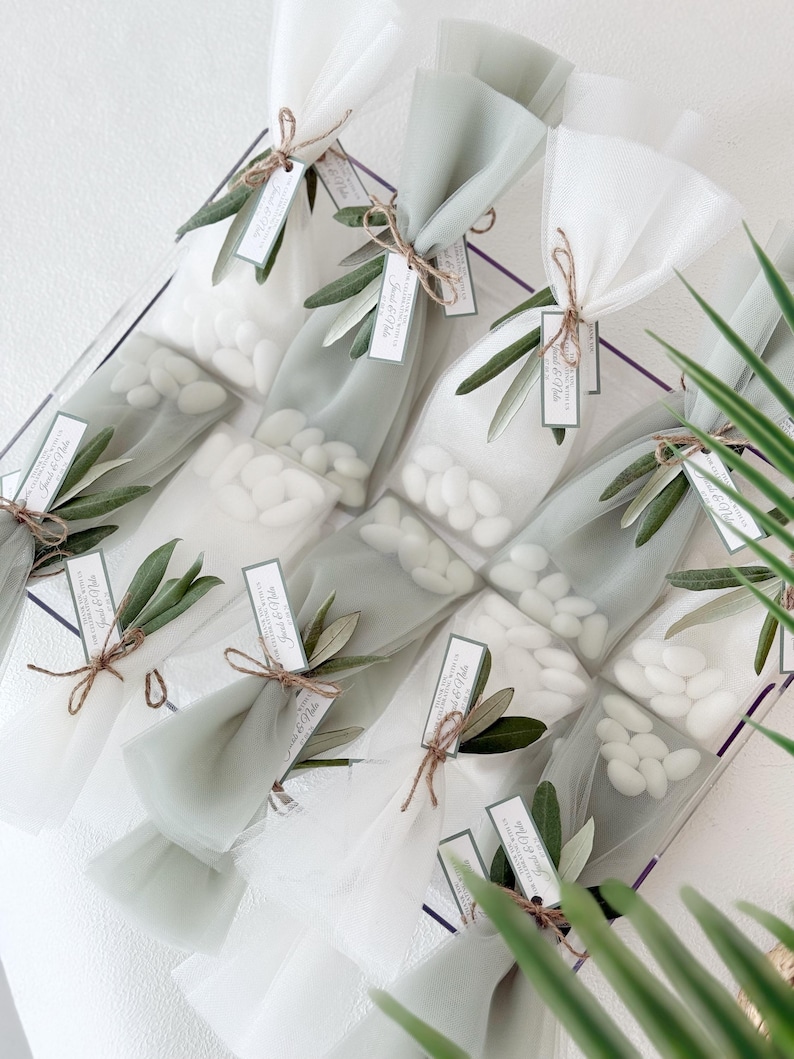 May include: Clear cellophane bags filled with white candies, tied with twine and adorned with olive branches and tags. The bags are arranged in a clear container, creating a visually appealing presentation. A perfect gift for a wedding or special event.