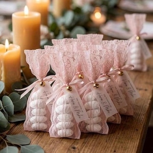 Pode incluir: Várias pequenas sacolas de renda rosa cheias de doces brancos são dispostas sobre uma mesa de madeira. Cada sacola é amarrada com uma fita rosa e um pequeno sino dourado. Etiquetas com texto pendem das fitas. Velas acesas e vegetação ao fundo.