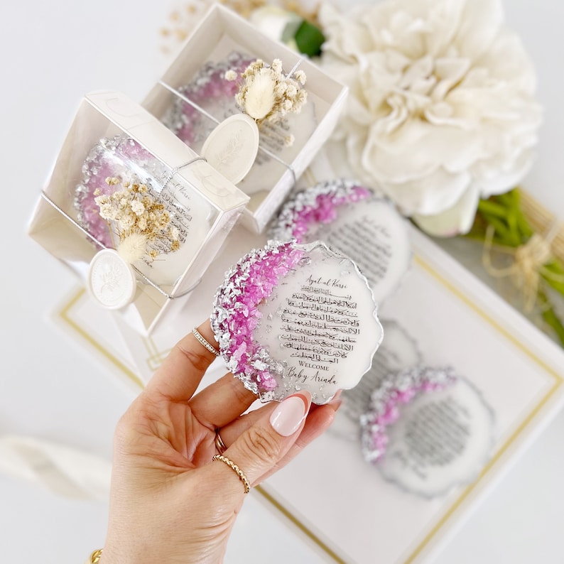 May include: A white and silver round resin keepsake with pink glitter and Arabic calligraphy. The text reads "Surah al-Kafirun" and "Welcome Baby Arida".