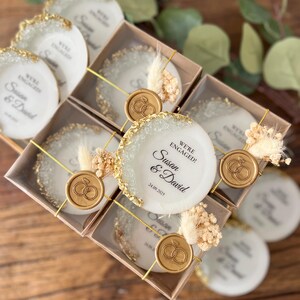 May include: Set of small white ceramic coasters with gold glitter edges and a gold wax seal with a ring design. Each coaster has the text "We're Engaged! Susan & David 24.09.2025" printed on it.