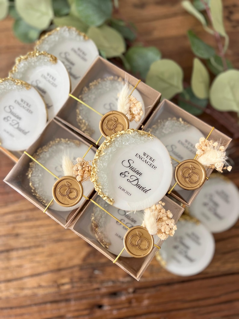 May include: Set of small white ceramic coasters with gold glitter edges and a gold wax seal with a ring design. Each coaster has the text "We're Engaged! Susan & David 24.09.2025" printed on it.
