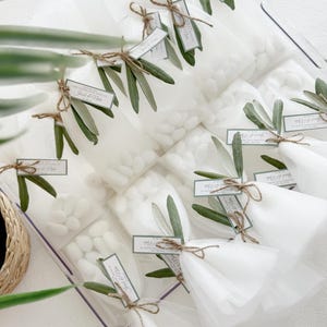 May include: Clear cellophane bags filled with white candies, tied with twine and adorned with olive branches and tags. The tags read "Thank you" and names. A decorative and elegant presentation for a special event.
