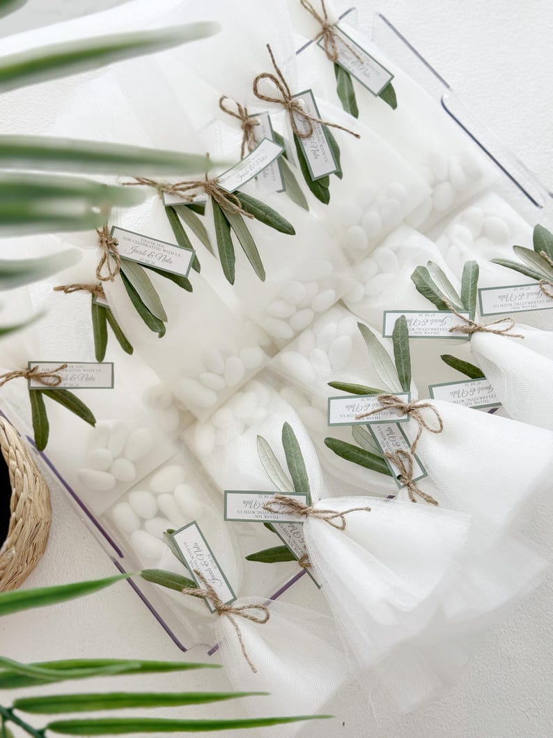 May include: Clear cellophane bags filled with white candies, tied with twine and adorned with olive branches and tags. The tags read "Thank you" and names. A decorative and elegant presentation for a special event.