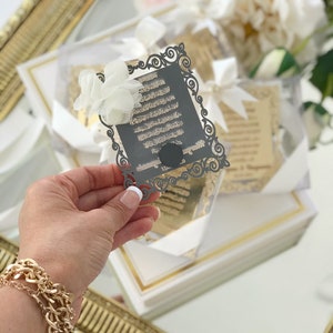 May include: A black and gold decorative card with Arabic calligraphy in a white frame. The card is held in a hand with a gold bracelet.