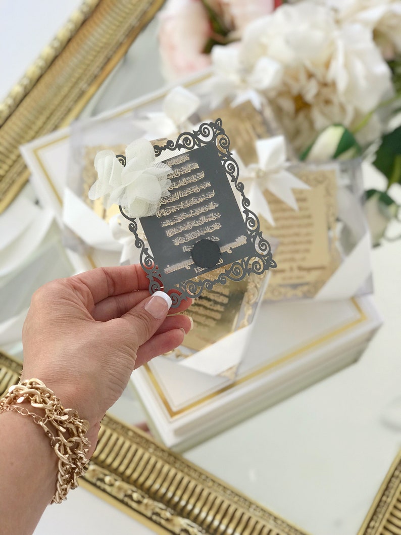 May include: A black and gold decorative card with Arabic calligraphy in a white frame. The card is held in a hand with a gold bracelet.