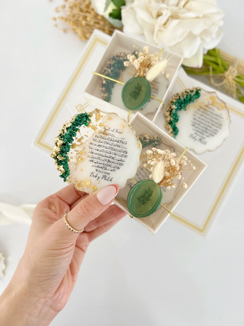 May include: A white, round, resin coaster with gold leaf and green accents. The coaster features Arabic calligraphy and a green wax seal with a leaf design. The text reads "Ayat al-Kursi" and "Welcome Baby Malik".