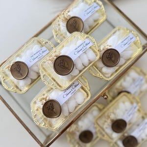 Könnte beinhalten: Eine Glasbox enthält mehrere kleine, goldfarbene Tabletts, die mit weißen Süßigkeiten gefüllt sind. Jedes Tablett ist in einem transparenten Material verpackt und mit einem braunen Wachsstempel versiegelt. Der Stempel zeigt ein botanisches Design und ein Band mit Text.