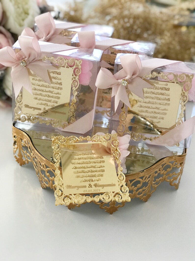 May include: Gold-colored decorative boxes with pink bows and a clear top. Each box contains a gold-colored plaque with Arabic calligraphy and the names "Maryam & Benjamin" and the date "05.05.2021".