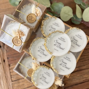 May include: White round favor boxes with gold glitter edges and a gold wax seal. The boxes have a white label with black text that reads "We're Engaged! Susan & David 24.09.2025".