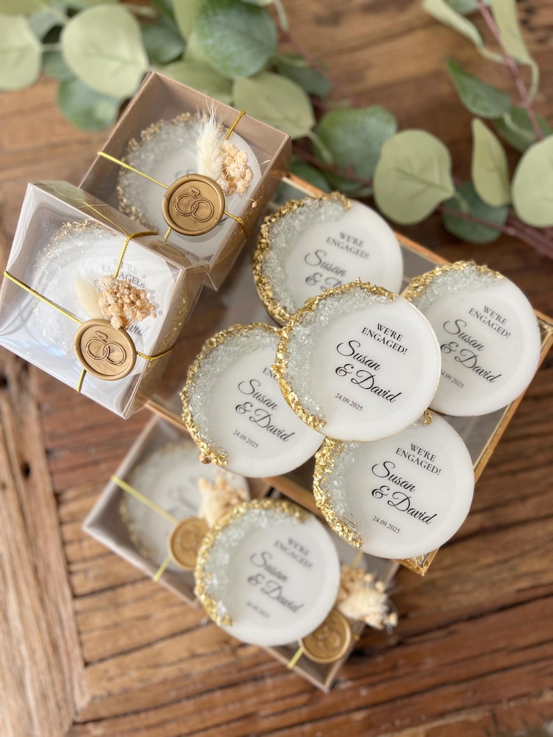 May include: White round favor boxes with gold glitter edges and a gold wax seal. The boxes have a white label with black text that reads "We're Engaged! Susan & David 24.09.2025".