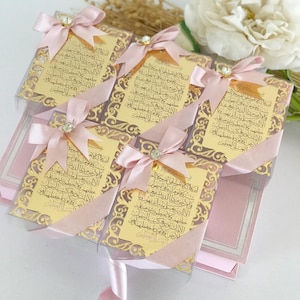 May include: Five small pink gift boxes with gold-colored decorative trim and a gold-colored inscription in Arabic. Each box is tied with a pink ribbon and a small rhinestone.