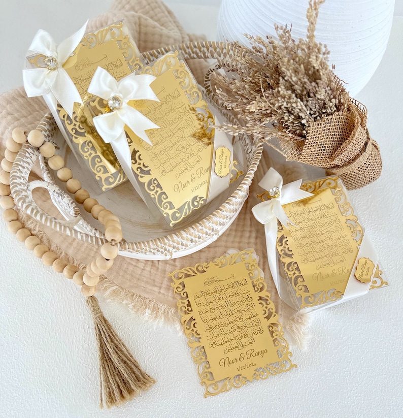 May include: Gold-colored decorative plaques with Arabic calligraphy and a white ribbon with a small diamond-shaped embellishment. The plaques are in clear plastic boxes and have the names "Neer & Ranya" and the date "1/32/2024" printed on them.