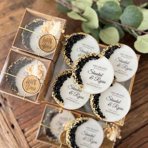 May include: White round save the date cards with black and gold glitter accents. The cards feature the text "Save the Date" and the names "Shantal & Ryan" with the date and location printed below. The cards are packaged in clear boxes with a gold wax seal that says "With Love".