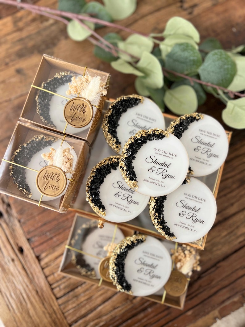 May include: White round save the date cards with black and gold glitter accents. The cards feature the text "Save the Date" and the names "Shantal & Ryan" with the date and location printed below. The cards are packaged in clear boxes with a gold wax seal that says "With Love".