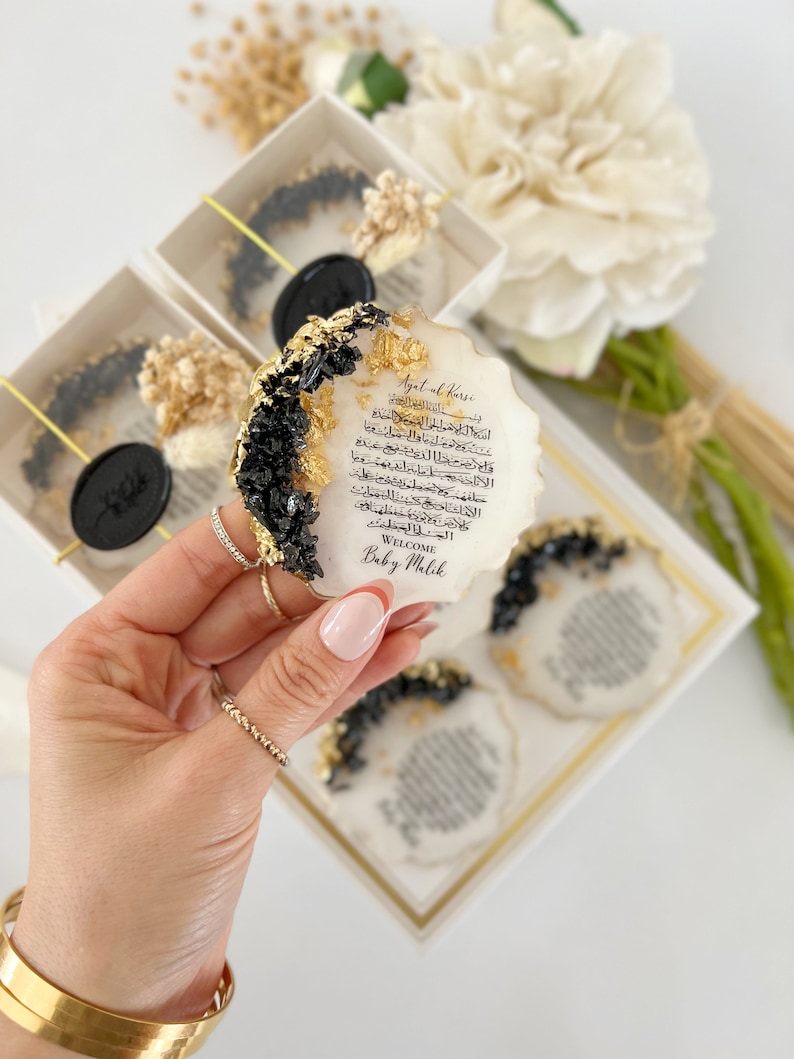 May include: A white oval resin plaque with black and gold accents. The plaque features Arabic calligraphy and the text "Ayatul Kursi" and "Welcome Baby Malik".