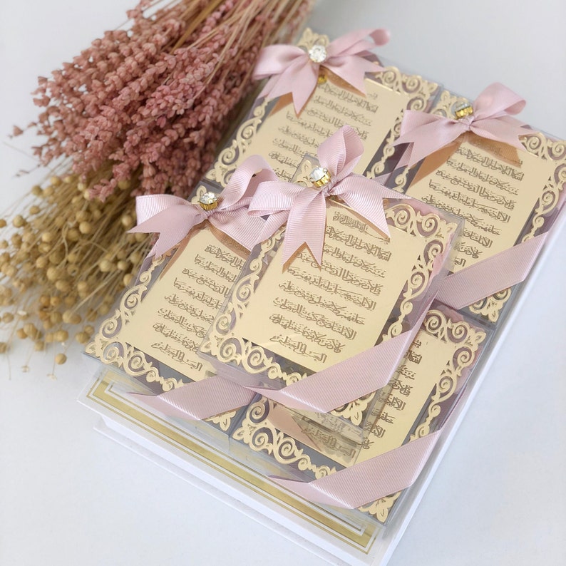 May include: A stack of small, square boxes with gold-colored decorative frames and Arabic text. Each box is tied with a pink ribbon and has a small, clear jewel in the center of the ribbon. The boxes are sitting on a white surface.