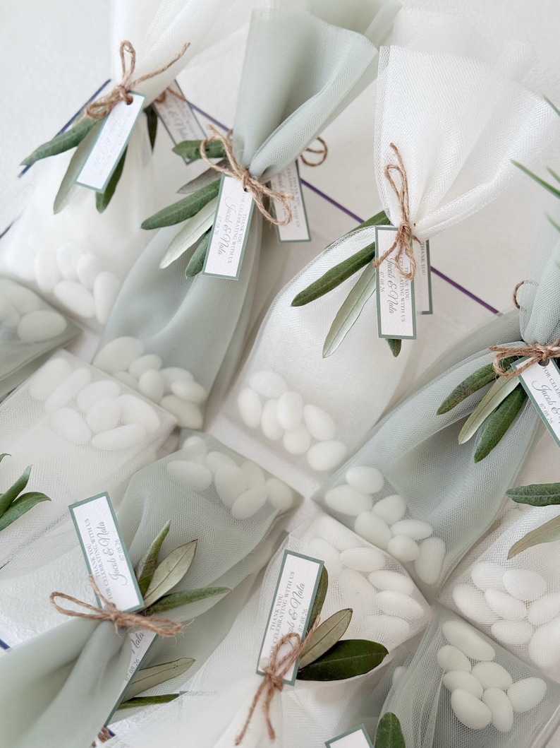 May include: Several small, clear bags filled with white candies, tied with twine and adorned with olive branches. Each bag has a small tag with text. The bags are arranged on a white surface, creating a delicate and elegant presentation.