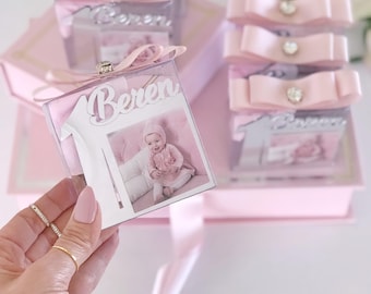 Puede incluir: Una caja de regalo cuadrada transparente con un lazo rosa y un adorno de pedrería. La caja contiene una foto de un bebé con ropa rosa. El nombre "Beren" está recortado encima de la foto. Cajas adicionales con lazos están apiladas en el fondo.