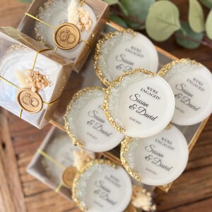 May include: White round favor boxes with gold glitter edges and a gold wax seal. The boxes have a printed message that says "We're Engaged! Susan & David 24.09.2025".