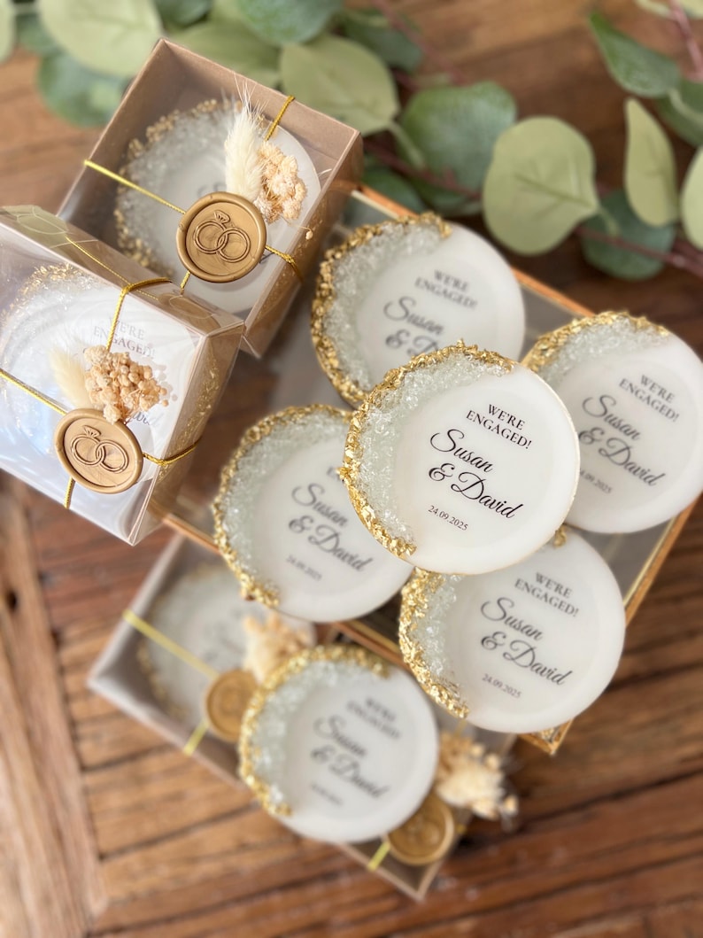 May include: White round favor boxes with gold glitter edges and a gold wax seal. The boxes have a printed message that says "We're Engaged! Susan & David 24.09.2025".
