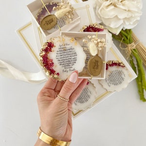 May include: A set of four round resin coasters with gold accents and dried flowers. Each coaster features a white background with Arabic calligraphy and a gold wax seal. The text on the coasters reads "Ayat al Kursi" and "Welcome Baby Arada".