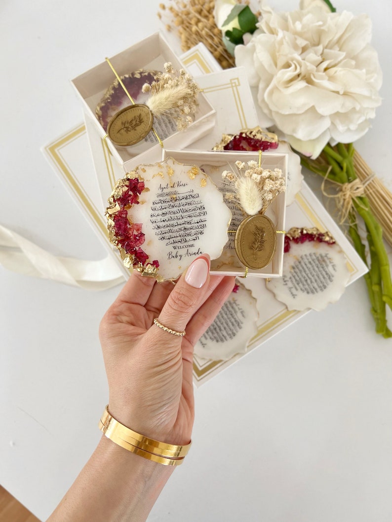 May include: A set of four round resin coasters with gold accents and dried flowers. Each coaster features a white background with Arabic calligraphy and a gold wax seal. The text on the coasters reads "Ayat al Kursi" and "Welcome Baby Arada".