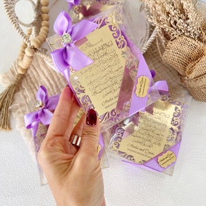 May include: Purple gift boxes with gold-colored decorative cutouts and Arabic calligraphy. The boxes are tied with purple ribbons and have a small gold tag with the names "Areeba and Qasim" and the text "Ramadan Mubarak 2021".
