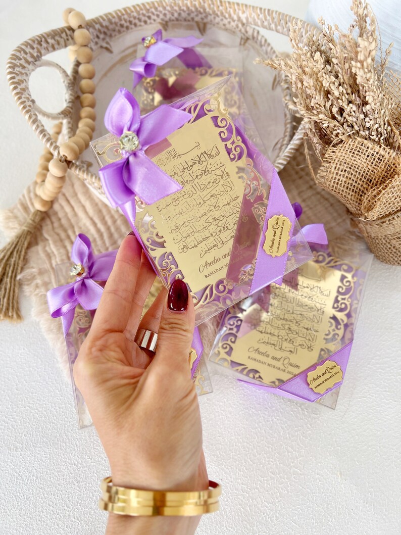 May include: Purple gift boxes with gold-colored decorative cutouts and Arabic calligraphy. The boxes are tied with purple ribbons and have a small gold tag with the names "Areeba and Qasim" and the text "Ramadan Mubarak 2021".
