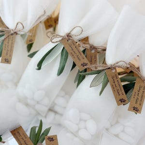 May include: Close-up of several small white tulle bags filled with white candies. Each bag is tied with twine and adorned with a sprig of green leaves and a small brown tag that reads "The Wedding of Zachary and Natalie 09.08.26".
