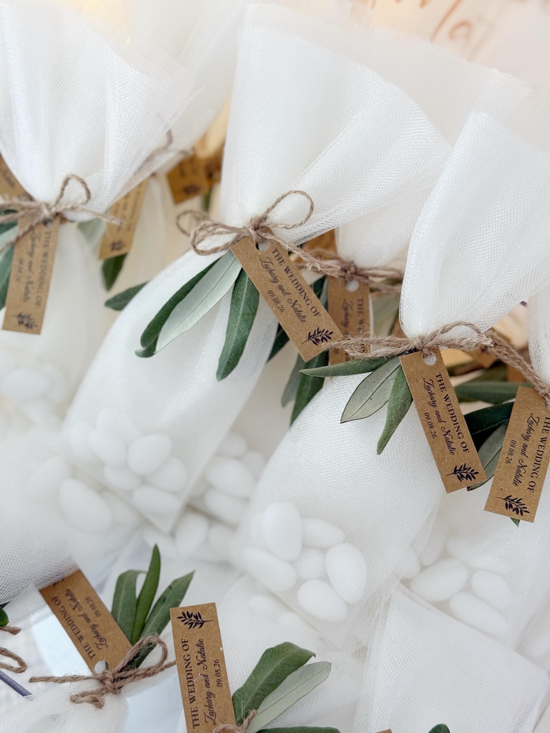 May include: Close-up of several small white tulle bags filled with white candies. Each bag is tied with twine and adorned with a sprig of green leaves and a small brown tag that reads "The Wedding of Zachary and Natalie 09.08.26".
