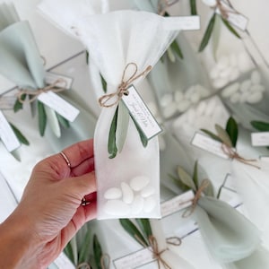 May include: Clear cellophane bags filled with white candies, tied with twine and adorned with olive branches. Each bag has a small tag with text. The bags are wrapped in white and light green tulle.