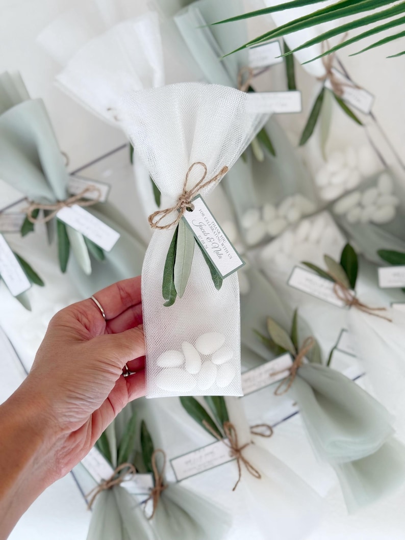 May include: Clear cellophane bags filled with white candies, tied with twine and adorned with olive branches. Each bag has a small tag with text. The bags are wrapped in white and light green tulle.