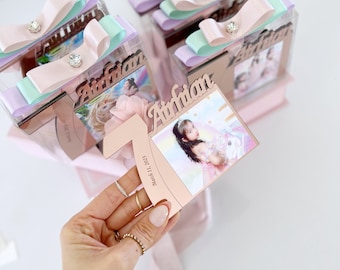 Puede incluir: Una caja de recuerdos de acrílico personalizada con un lazo de cinta pastel y un centro de diamantes de imitación. El frente muestra un recorte del número uno, el nombre Airfian, la fecha 11 de marzo de 2023 y una foto de un niño.