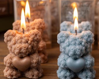 May include: Two bear-shaped candles, one tan and one light blue, each featuring a textured rose-like surface and holding a smooth heart in the center. Both candles are lit, with visible flames, and are positioned in front of clear packaging.