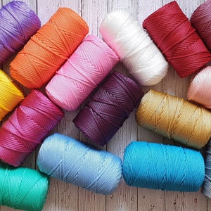 May include: An assortment of brightly coloured nylon cord spools, including shades of purple, orange, pink, white, red, and teal. The spools are arranged on a light wood-grain surface, displaying the vibrant colours and textures of the craft material.
