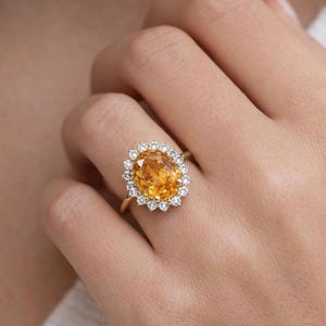 Puede incluir: Un anillo de oro con una gran gema amarilla de corte ovalado, rodeada de gemas más pequeñas y transparentes. El anillo está engastado en una delgada banda de oro, creando un diseño clásico y elegante. El anillo se muestra en una mano.