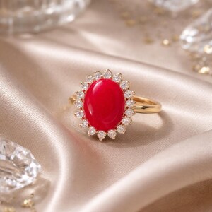 May include: A gold ring with a large, oval, bright red gemstone encircled by a halo of small, clear gemstones. The ring is displayed on a satin surface, reflecting light. The ring's band is a simple, polished gold, creating a luxurious feel.