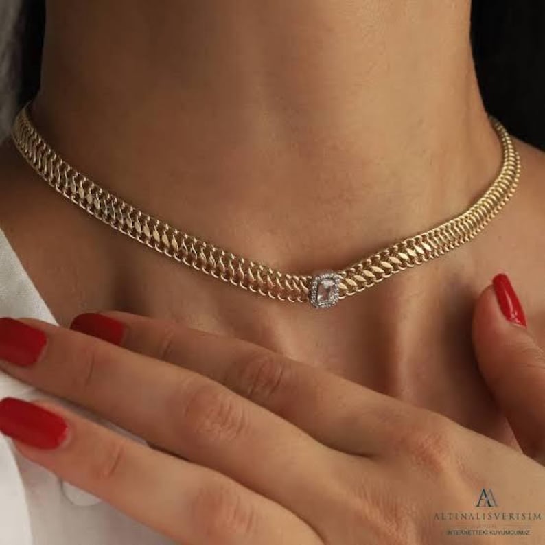 May include: A gold chain necklace with a square-shaped clear stone pendant. The necklace is on a model's neck.