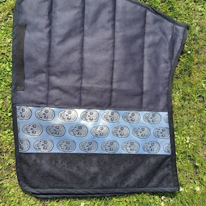 Könnte beinhalten: Eine schwarze Sattelunterlage mit Steppmuster. Ein hellblaues Band mit einem sich wiederholenden Totenkopfmuster ist mittig auf der Unterlage angebracht. Der untere Teil der Unterlage hat ein schwarzes Blumenmuster.