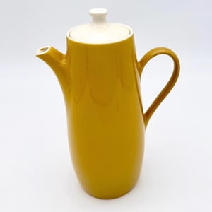 May include: Mustard yellow ceramic coffee pot with a white lid and handle. The pot has a curved body and a spout for pouring. The handle is a loop shape. The lid has a small knob.