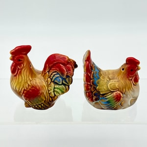 May include: A pair of ceramic salt and pepper shakers shaped like a rooster and a hen. The rooster features a red comb and wattle with brown, yellow, and blue plumage. The hen has a red comb with brown, yellow, and blue feathers on its wings.